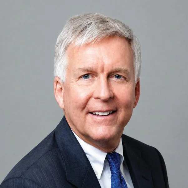 Professional headshot of a smiling, gray-haired older man in a dark suit, white shirt, and blue tie against a gray background.