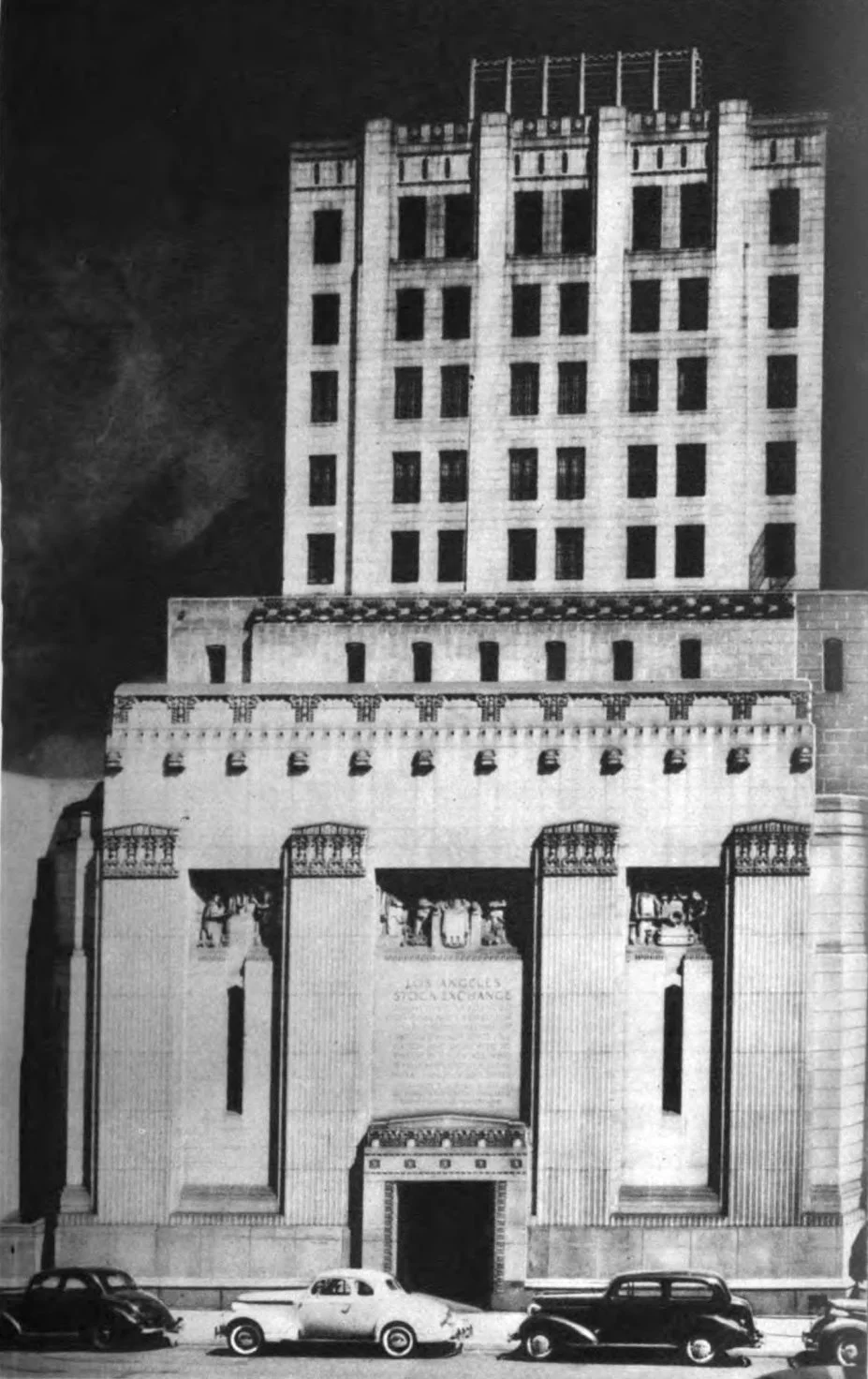 Black and white photo of a tall, multi-story building with intricate architectural details, fitting with classic early 20th-century style, along with several vintage cars parked in front of it.