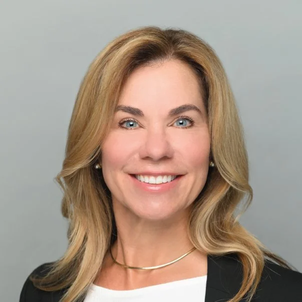 Professional headshot of a smiling blonde woman with blue eyes, wearing a black blazer and white top against a gray background.