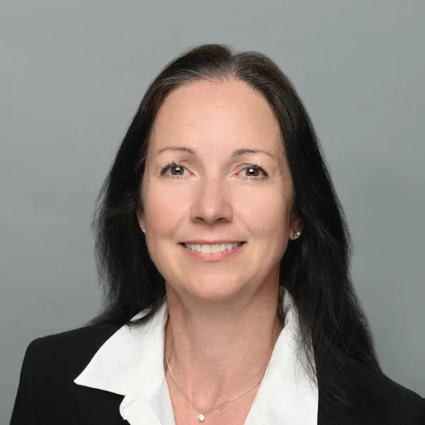 Professional headshot of a smiling woman with long dark hair in a black blazer and white shirt against a gray background.