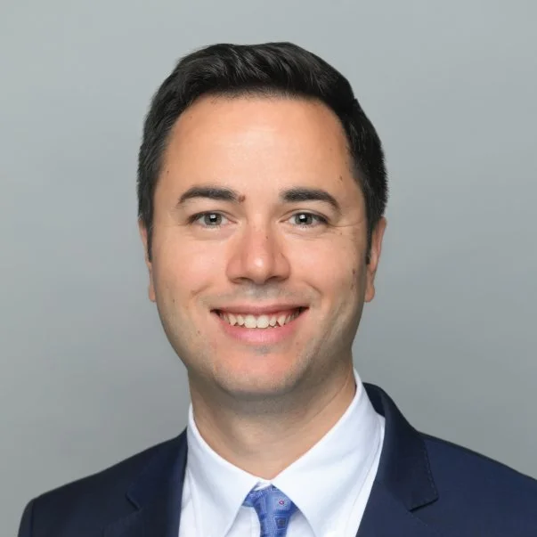 Professional headshot of a smiling man in a navy suit, white shirt, and patterned tie against a plain gray background.