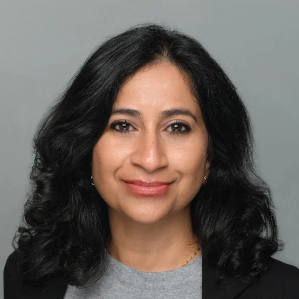 Professional headshot of a smiling woman with wavy black hair, wearing a black blazer and gray top against a gray background.