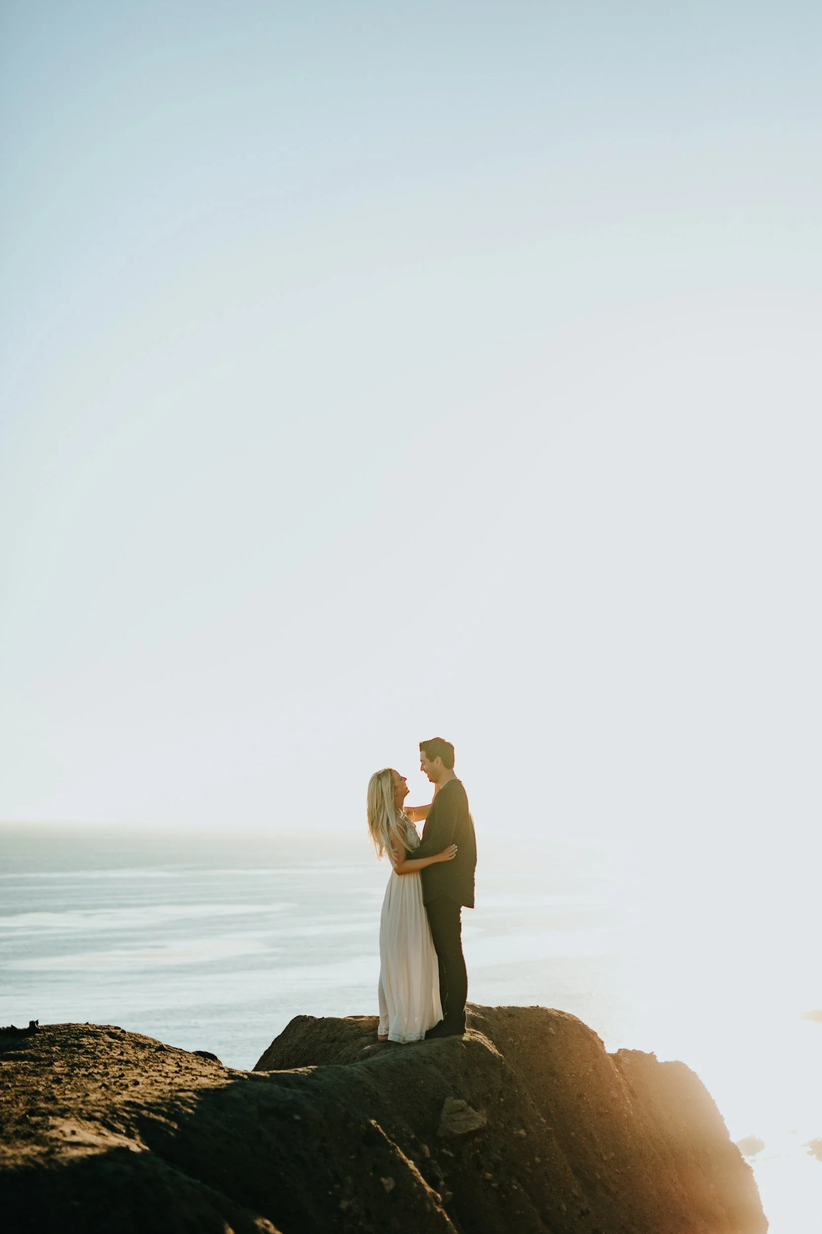 Couple embracing on a coastal rock at sunset during an engagement session, photographed by Snap Sync Studio.