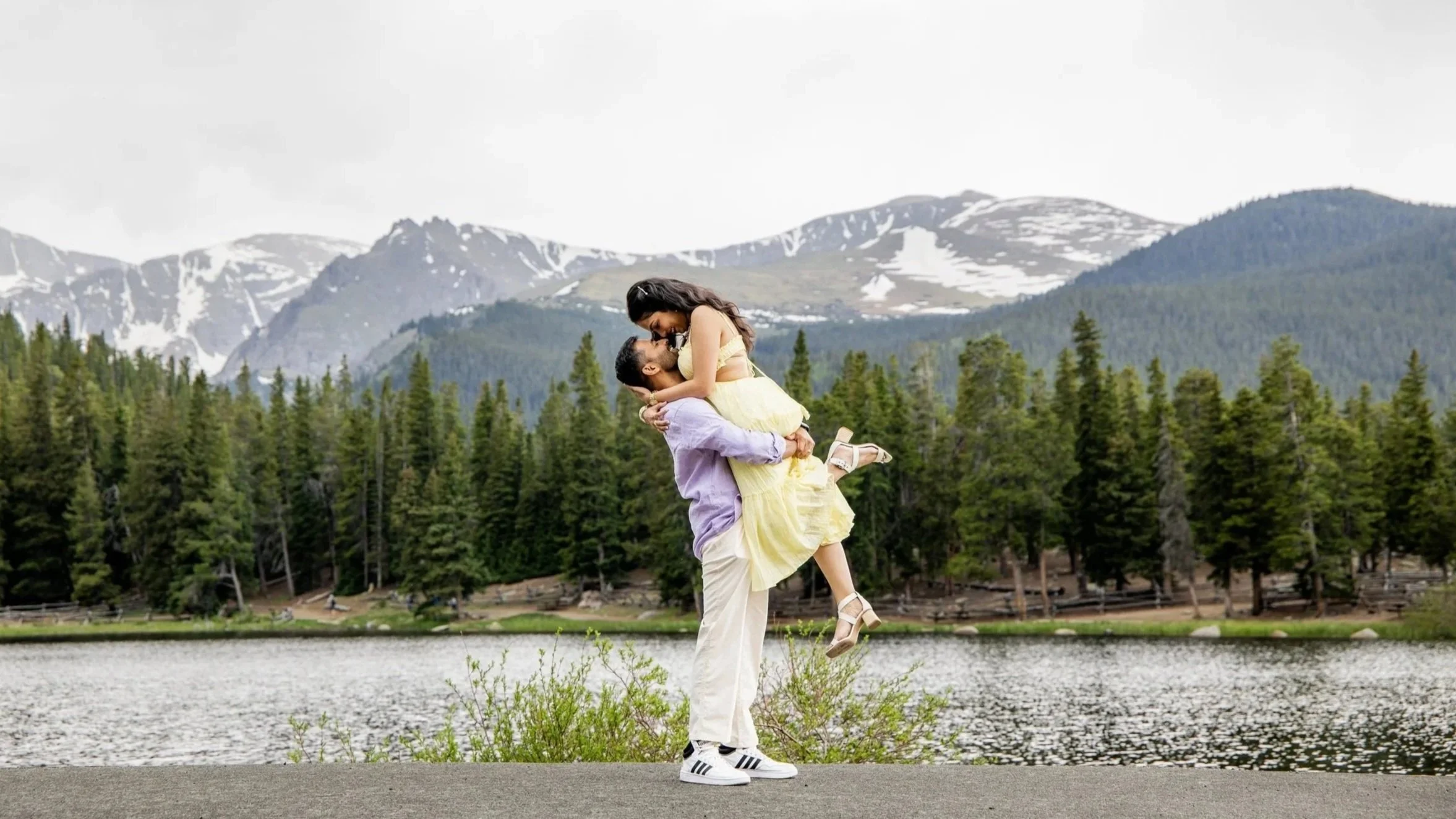 A couple outdoors in a mountainous area with snow-capped peaks, green trees, and a river. The man is holding the woman, who is leaning forwards with arms wraped around him, smiling in a yellow dress.