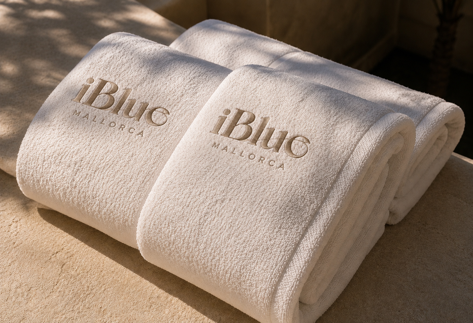Two folded white towels with the words 'iBlue Mallorca' embroidered on them, placed on a beige surface with shadows cast from nearby trees.