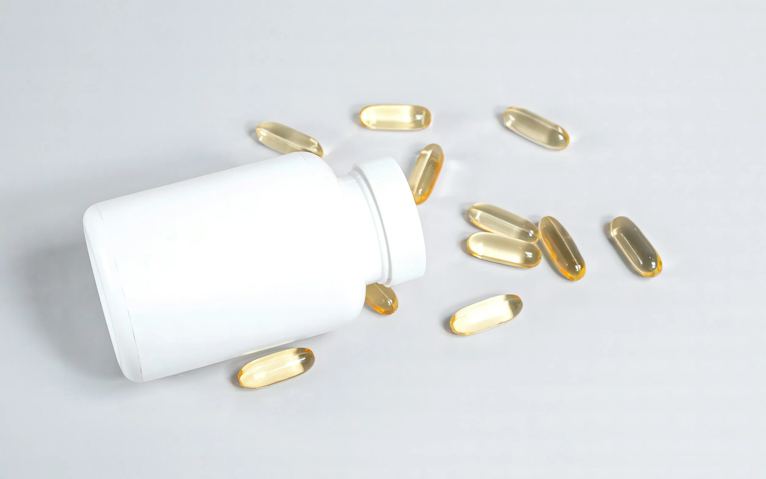 A white pill bottle lying on its side with several transparent, yellowish capsules spilled out onto a white surface.