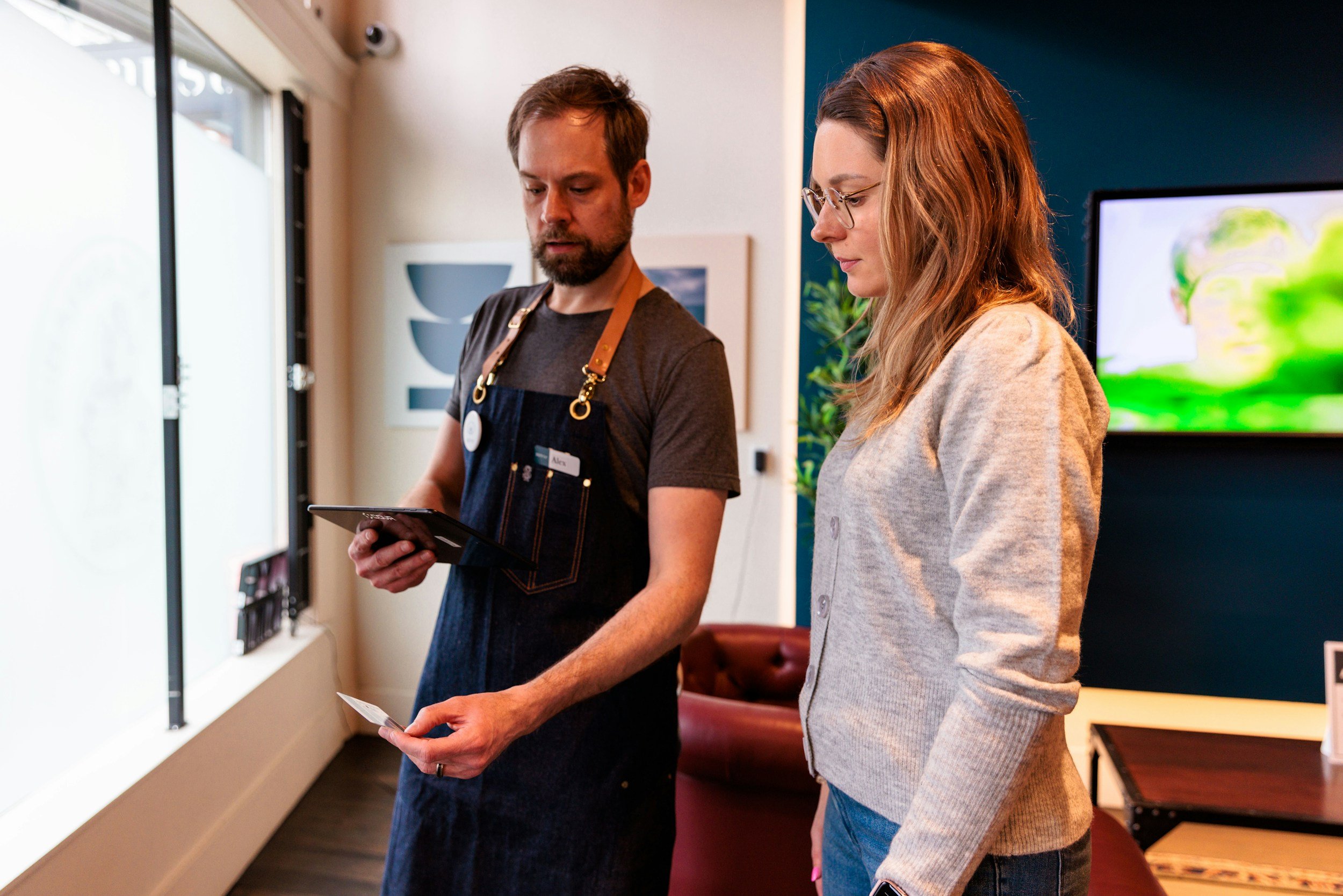 A man and a woman are standing inside a room with a large window. The man, wearing a dark shirt, apron, and name tag, is looking at a tablet. The woman, with long hair and glasses, is wearing a light gray cardigan and looking at the tablet too. There is a television screen displaying a colorful image in the background.