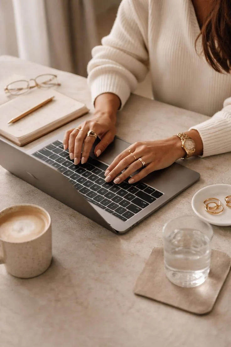 Une personne utilisant un ordinateur portable sur une table, avec une tasse de café, une petite assiette avec des bagues, un verre d'eau, un carnet, un stylo, et des lunettes. La personne porte une montre et des bijoux.