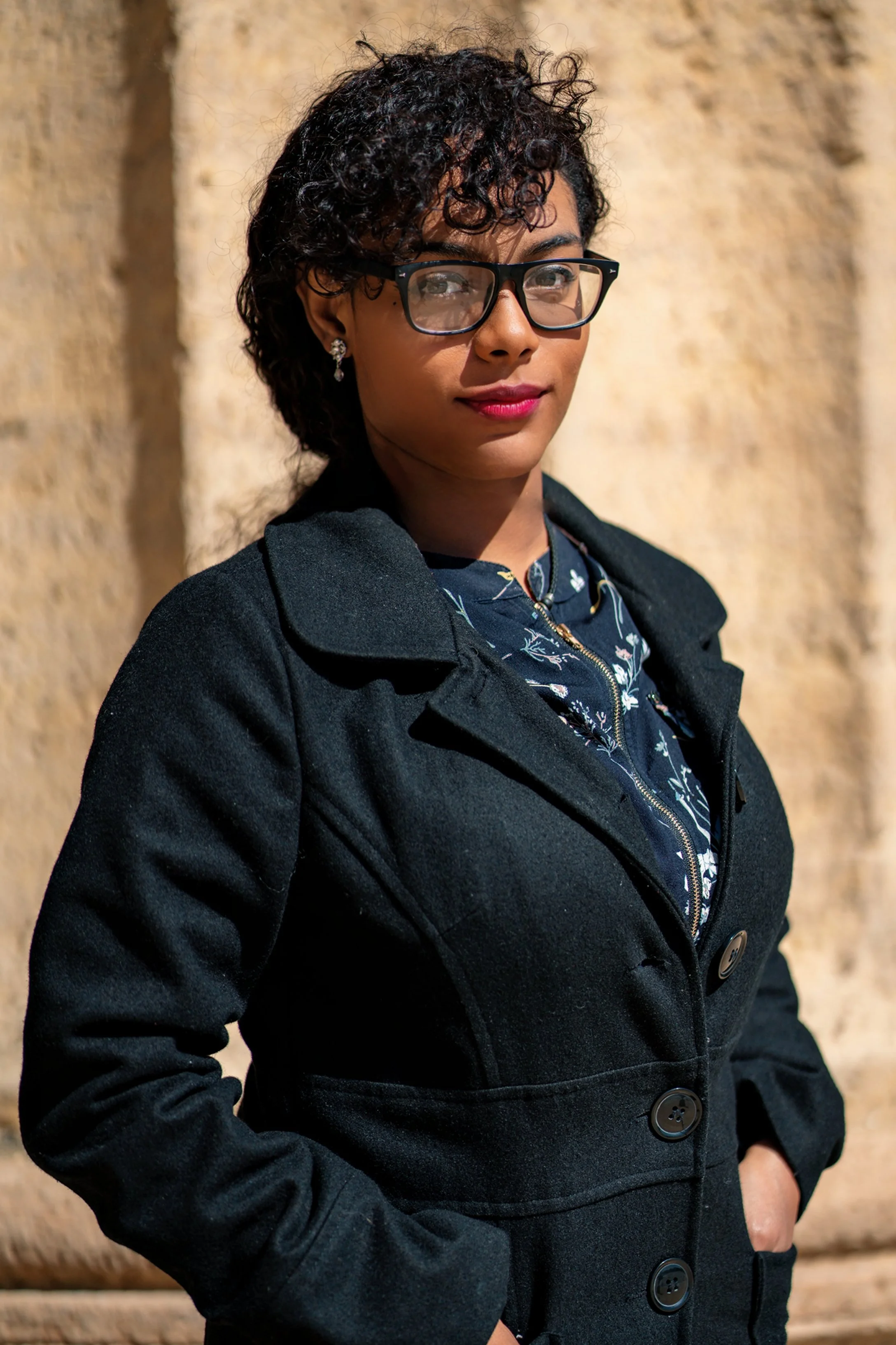 A woman wearing glasses, earrings, a navy shirt with floral print, and a black coat standing against a beige stone wall.