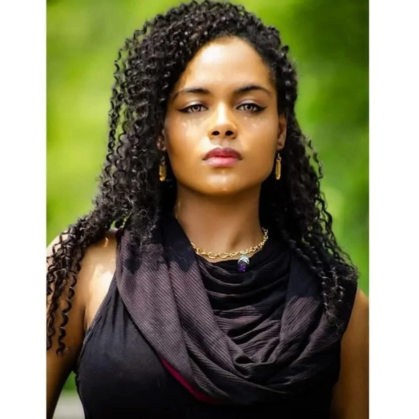 A woman with dark curly hair wearing a black top and a black scarf, gold jewelry, and standing outdoors with a blurred green background.