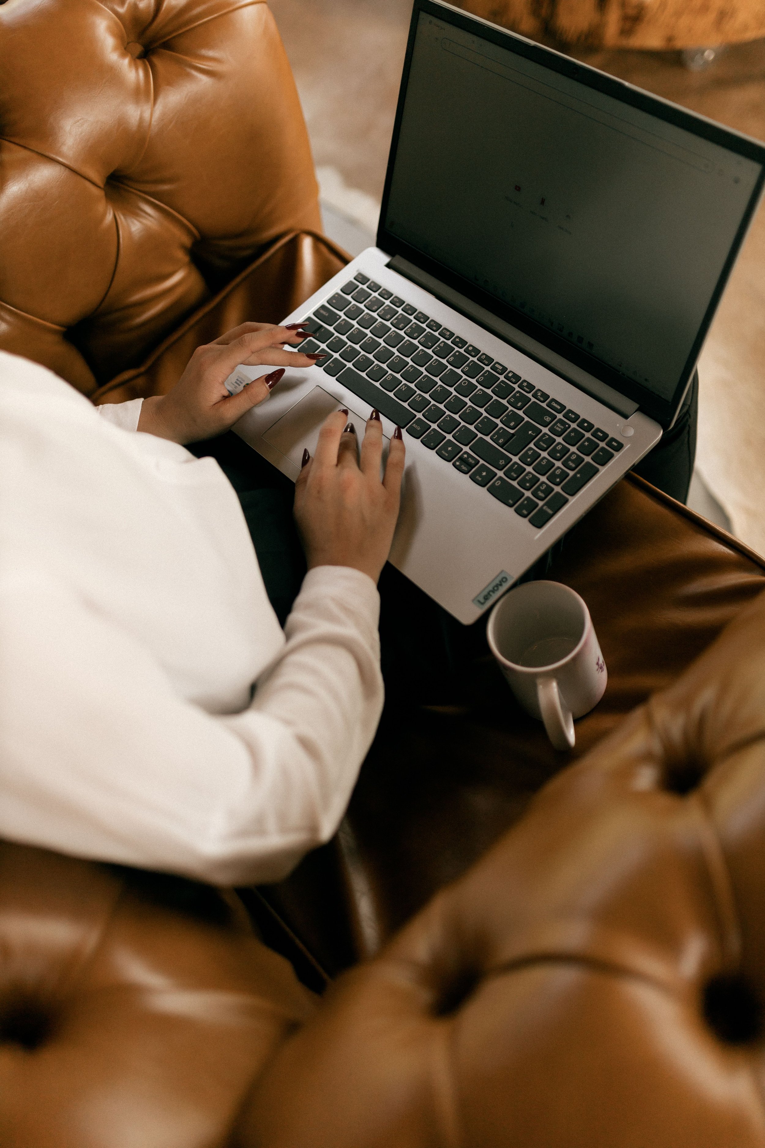 A person sitting on a brown leather tufted couch working on a silver laptop, wearing a white long sleeve shirt with manicured nails, next to a white coffee mug.