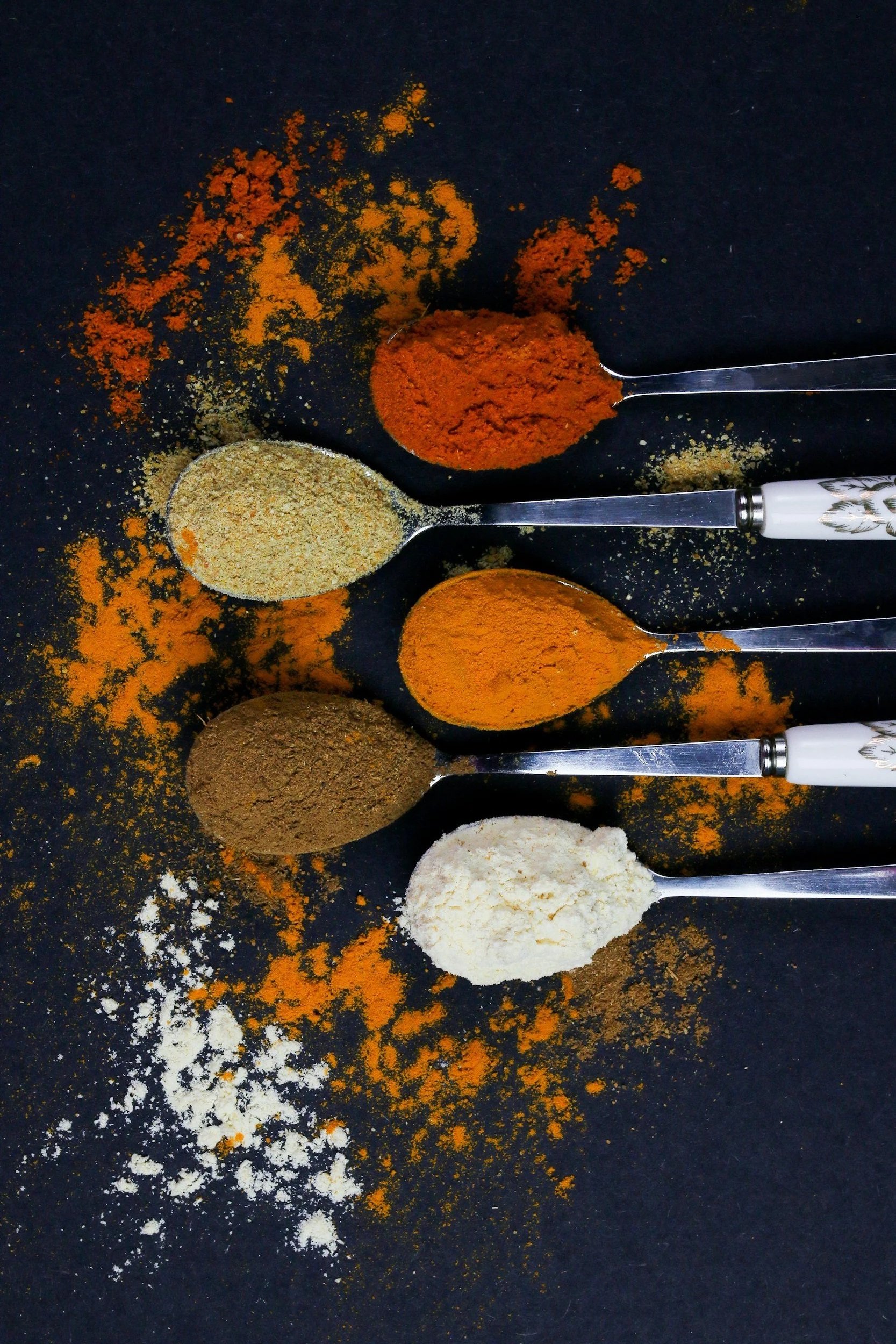 Six silver measuring spoons filled with different spices arranged on a black surface. The spices include white, orange, beige, brown, and yellow colors, with some spices spilled around the spoons.