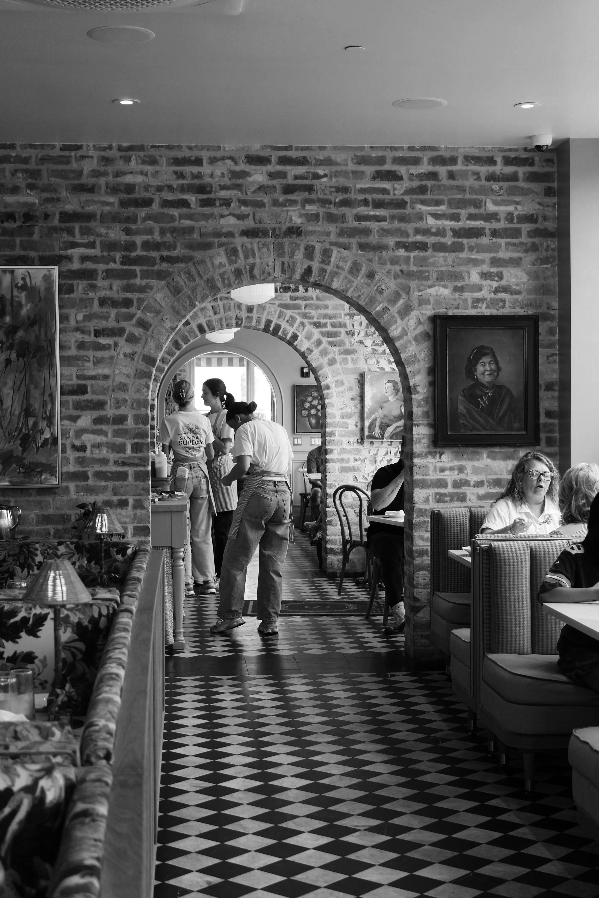A black and white photo of a restaurant interior with exposed brick walls and an arched doorway. People are standing and sitting, with some staff serving food. There are framed paintings hanging on the walls, and a checkered floor design.