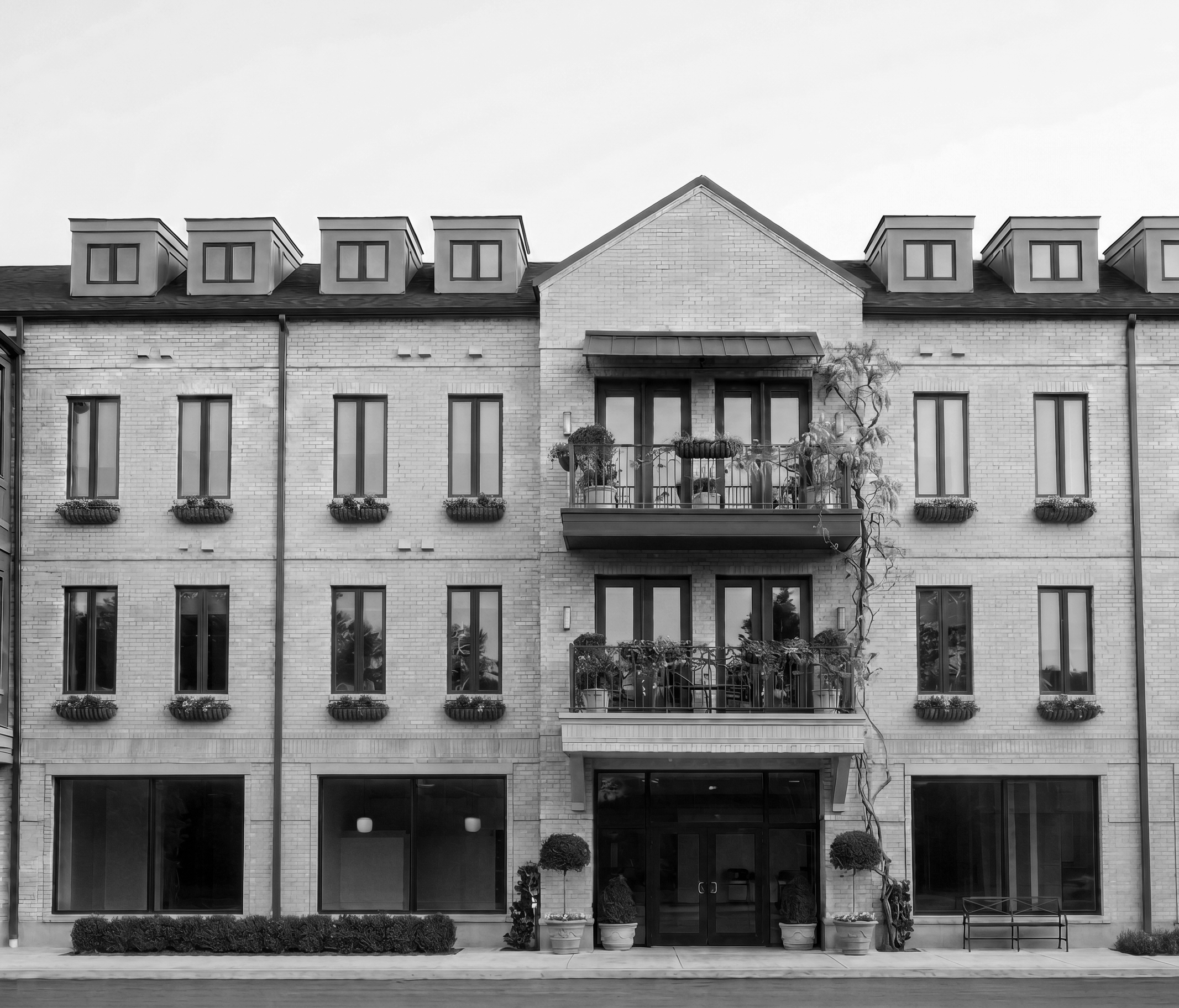 Black and white image of the front of a modern apartment building with multiple windows, some with flower boxes, and small balconies with potted plants.
