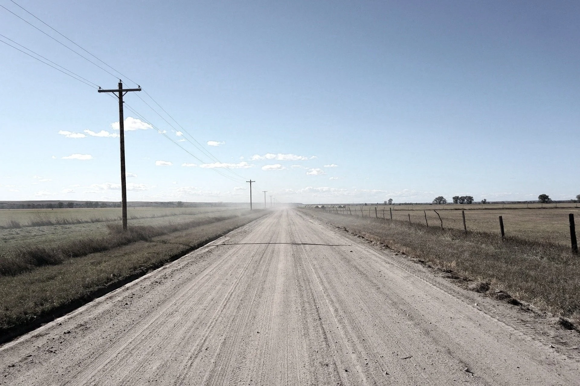 A straight dirt road runs through a rural landscape with grassy fields on either side. Utility poles with power lines stretch into the distance under a clear, blue sky with scattered small clouds.