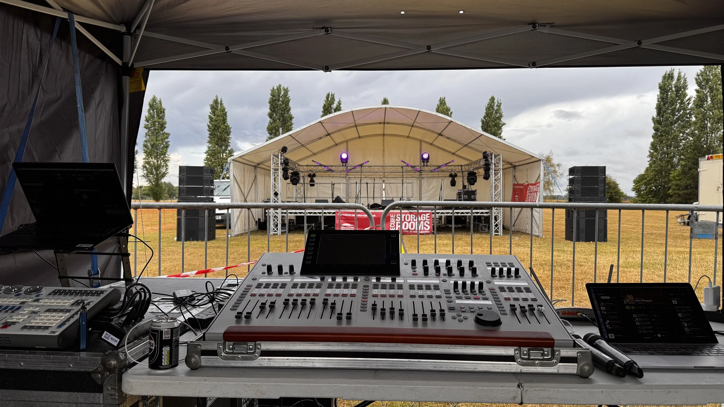 Music stage set up outdoors on a field with trees in the background, viewed from behind a DJ or sound technician's equipment including mixers and screens under a canopy.