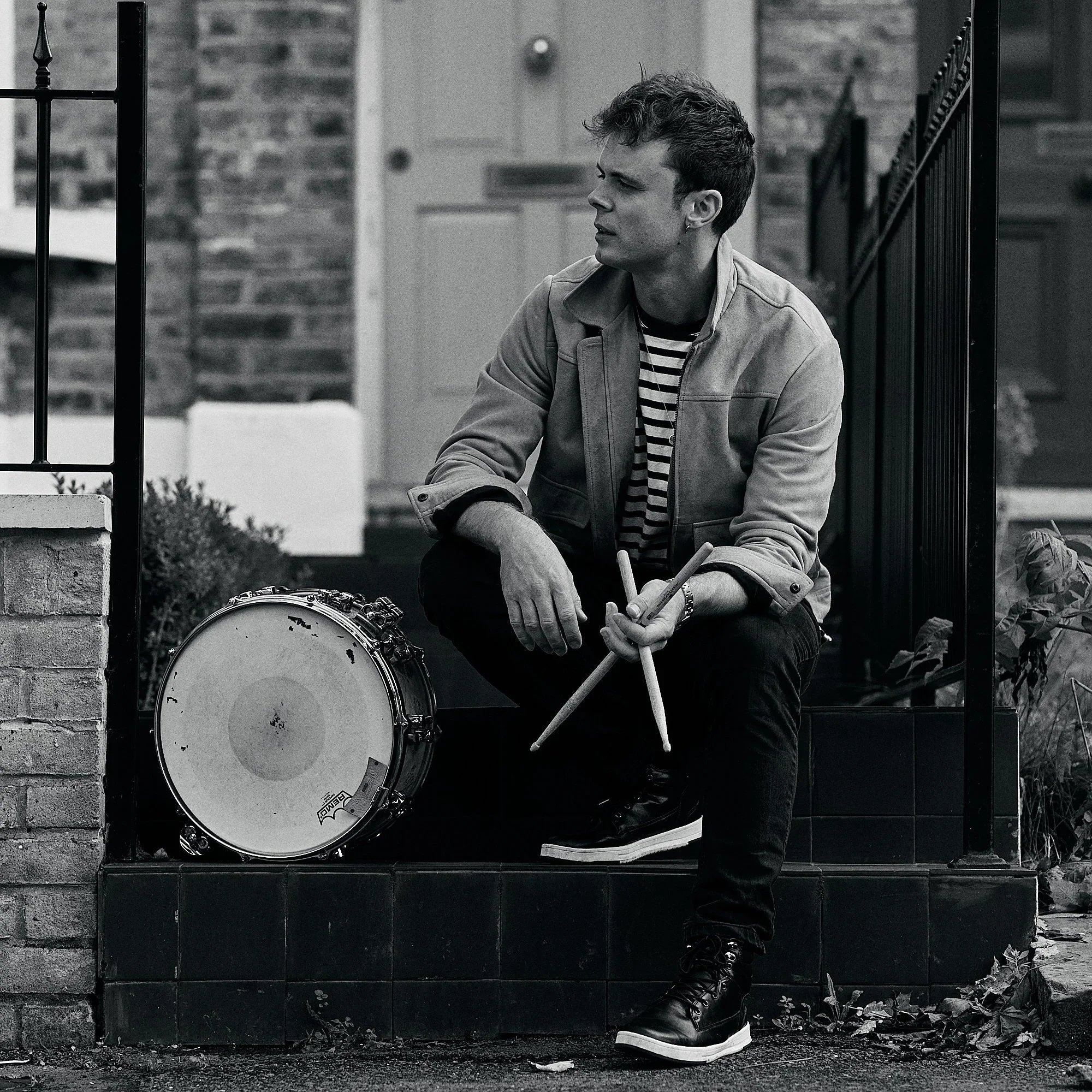 A man with curly hair, wearing a striped shirt under a jacket, sitting on a low wall next to a drum, looking to the side. The background shows a brick house with a door, a fence, and some plants and leaves on the ground. The photo is in black and white.