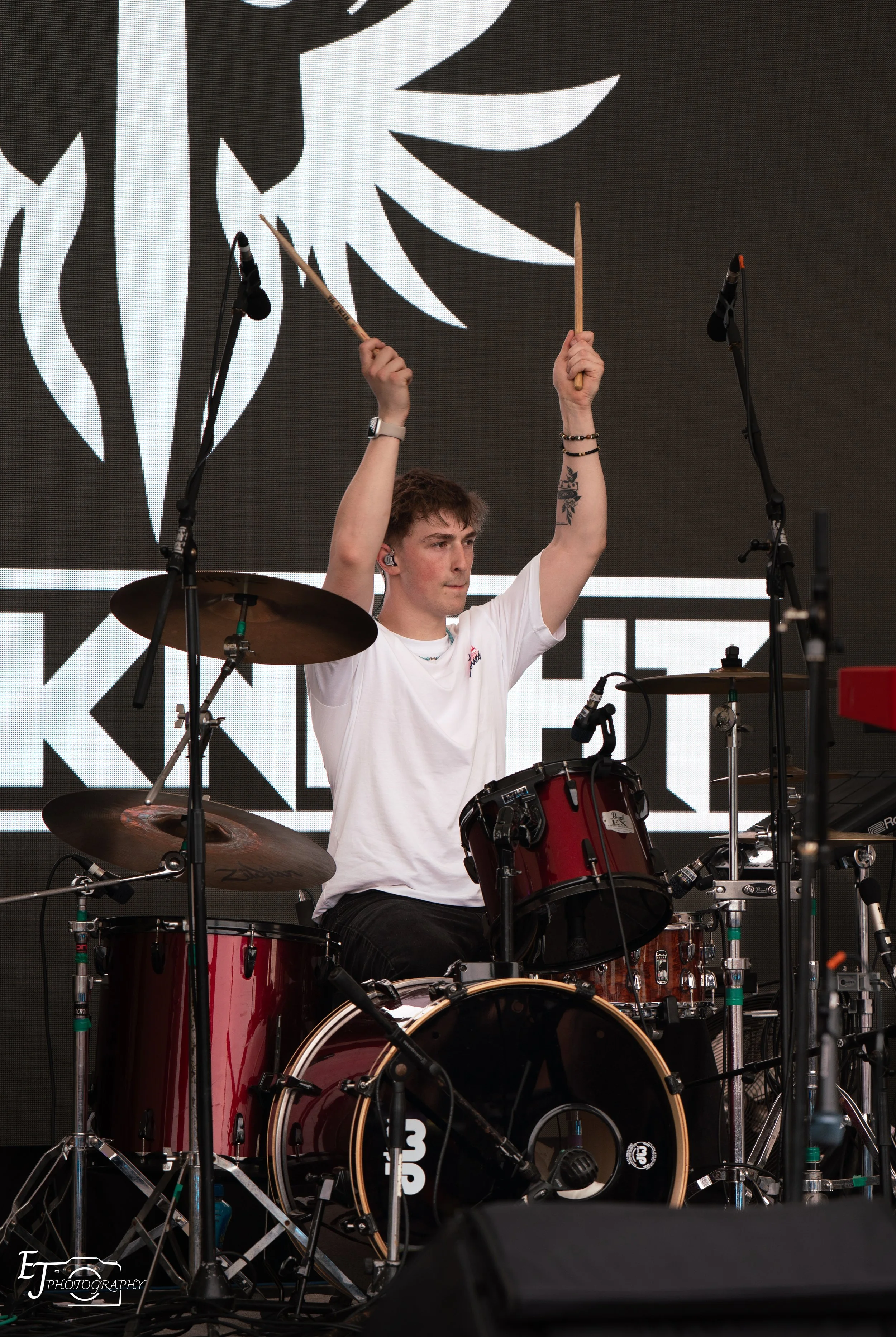 Oli Hyland is playing a red drum set on an outdoor stage with a large black and white graphic background. He is holding drumsticks, wearing a white t-shirt, and has tattoos and accessories on his arms.