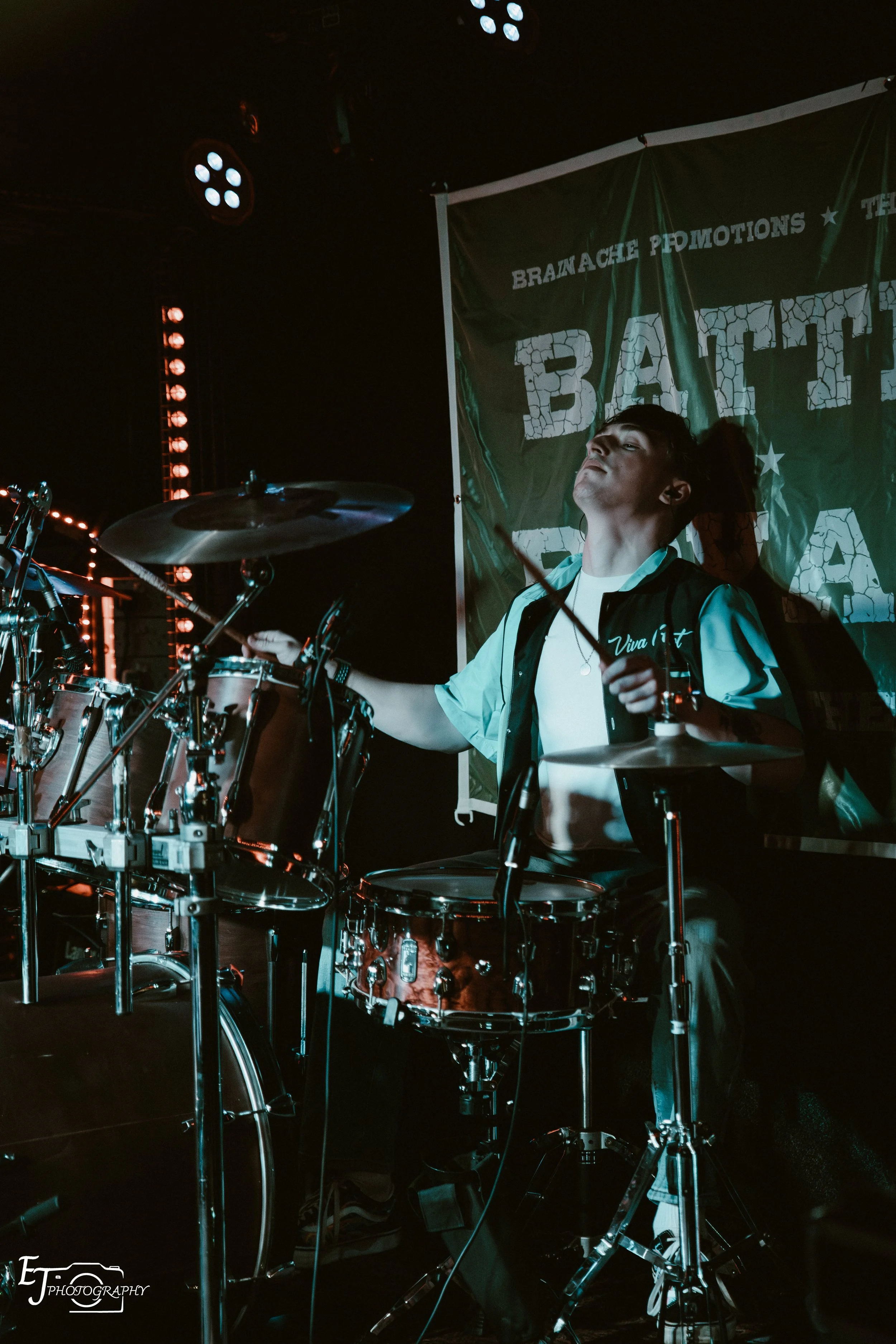 Oli Hyland is playing the drums on stage during a concert with stage lights and a banner in the background.