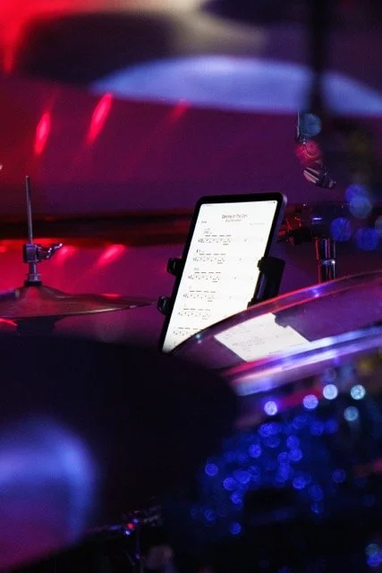 A tablet mounted on a stand on a drum set, displaying sheet music or notes, in a dimly lit room with red and blue stage lighting.