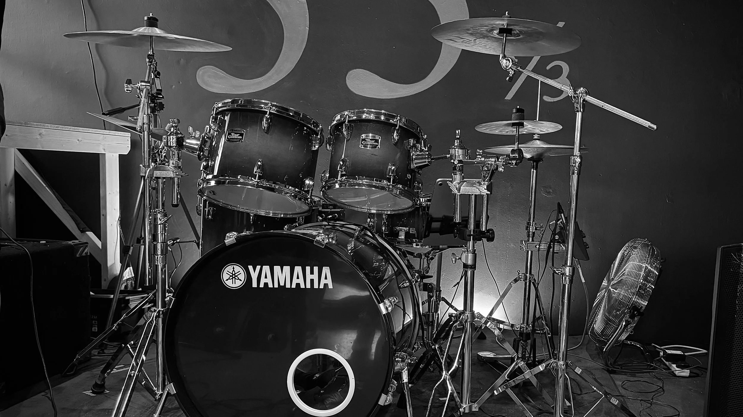 A black and white photograph of a Yamaha drum set on a stage with cymbals, drums, guitar pedals, a fan, and a wall with a large artistic design in the background.