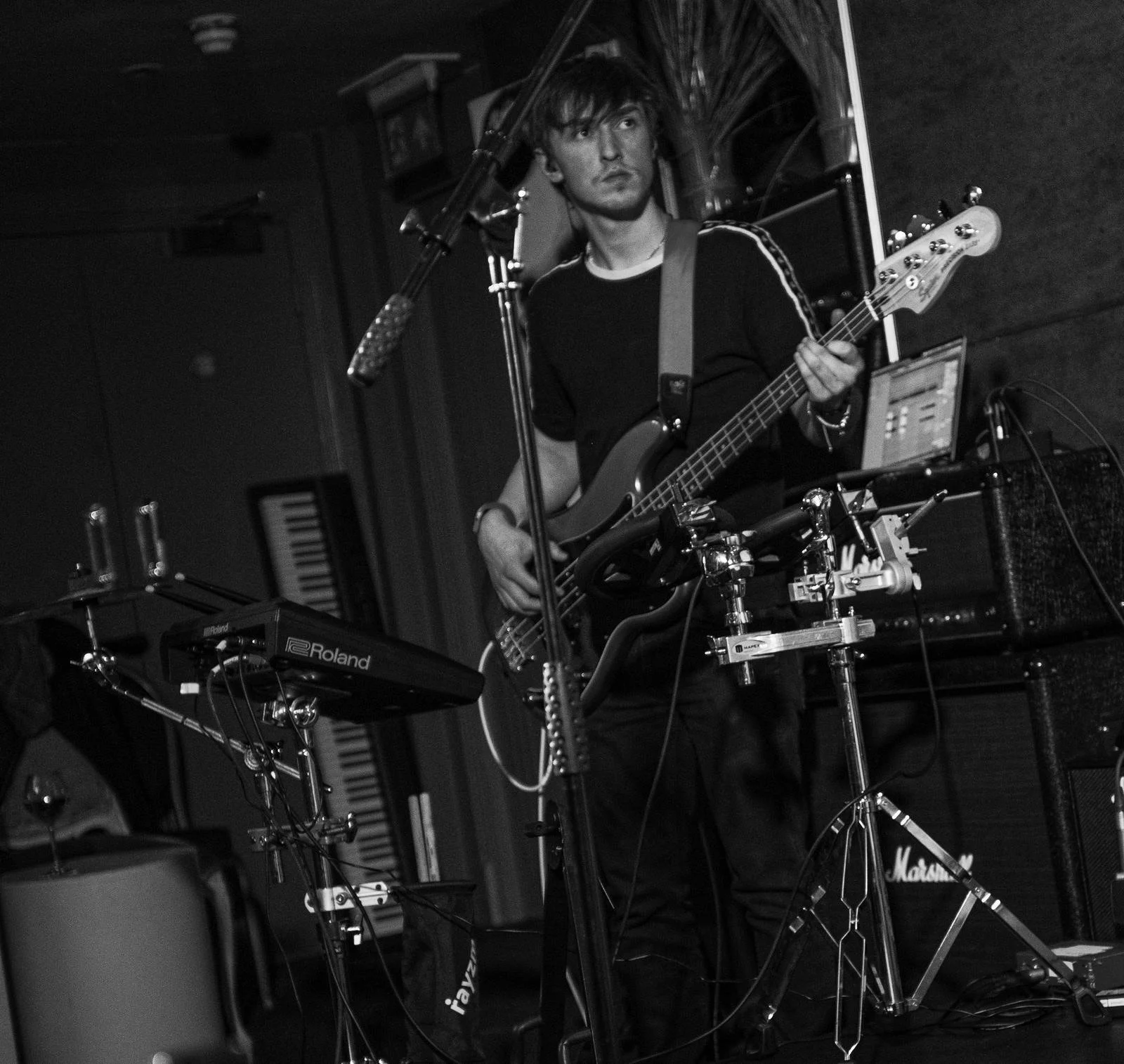 Oli Hyland is playing bass guitar at a live music venue, surrounded by electronic music equipment including a Roland sample-pad and Marshall amplifier, with microphones set up in front of him.