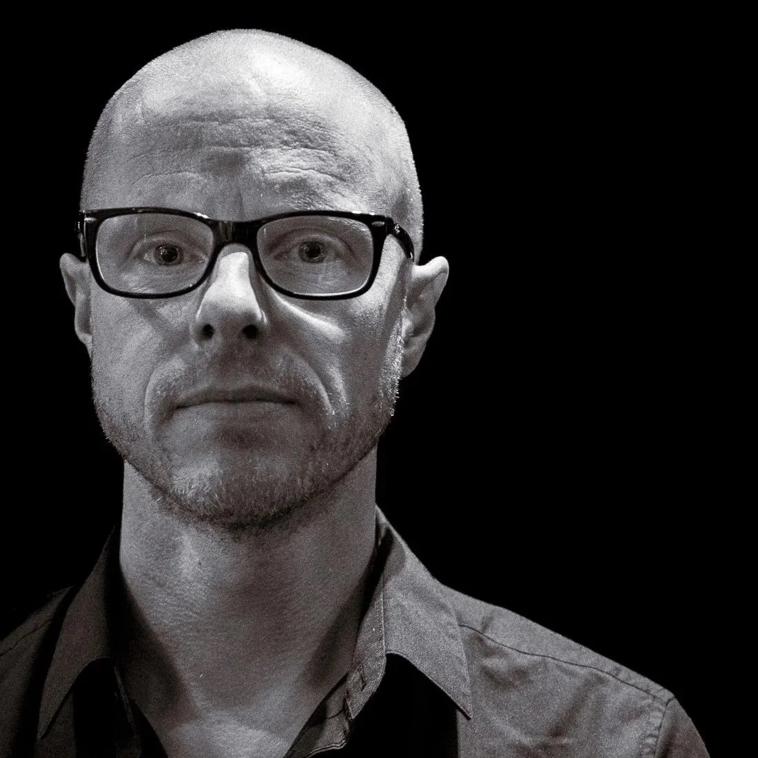 A black-and-white close-up portrait of a man with glasses, a bald head, and a slight beard, against a dark background.