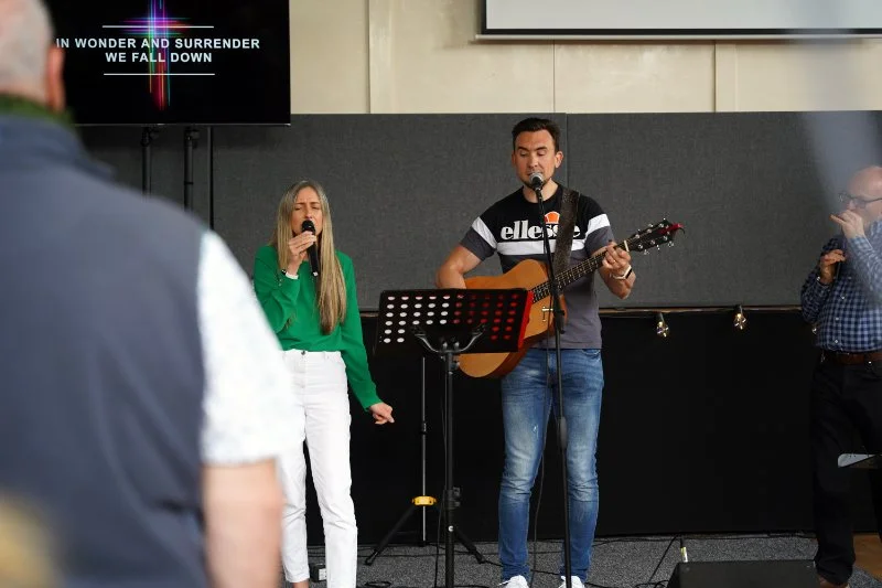 A man and a woman on stage in church singing and playing guitar