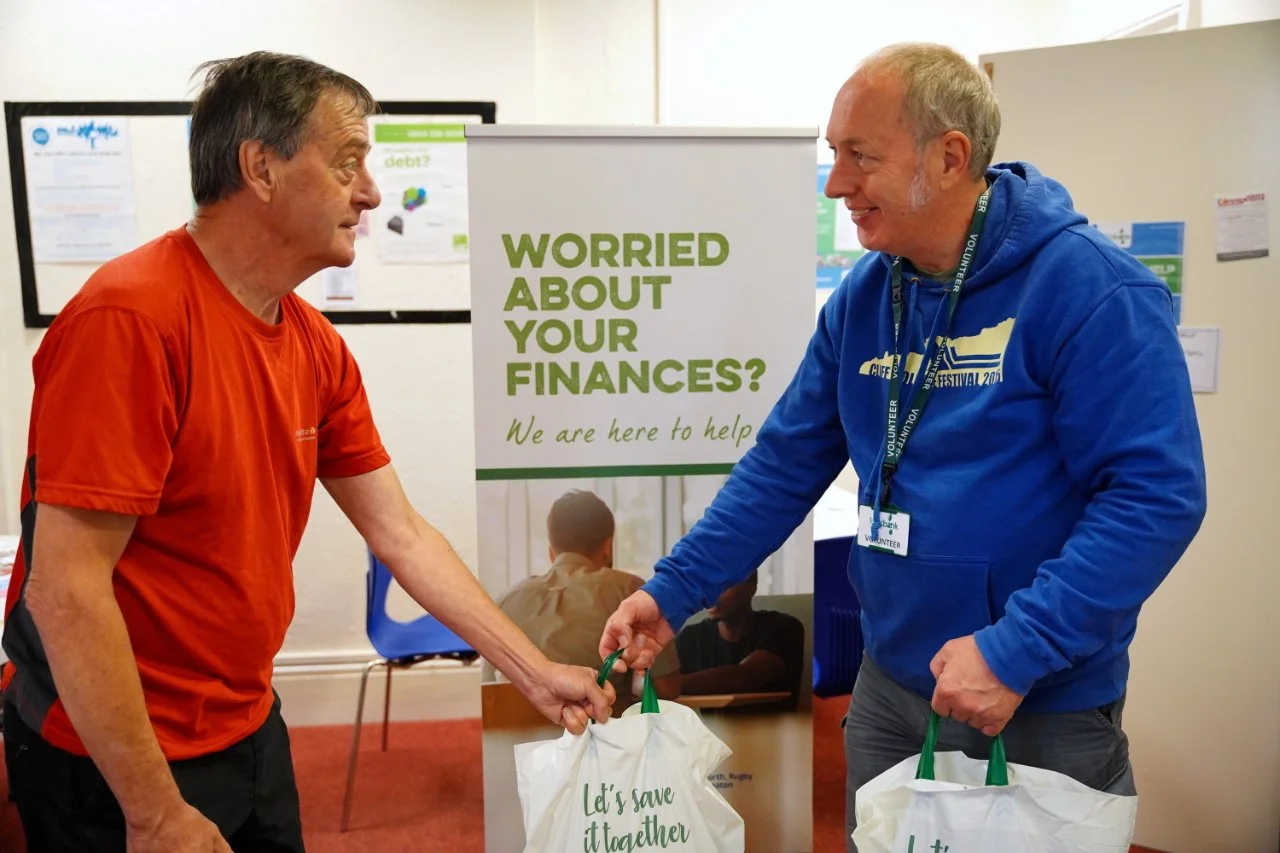 A Foodbank worker in a blue jumper handing over two bags of shopping to a client, standing in front of a roller banner showing the words 'Worred about your finances? We are here to help'
