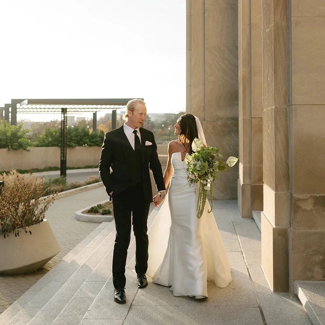 A newlywed couple walking hand in hand outside a building on a sunny day, with the bride holding a bouquet of white and green flowers. The groom is dressed in a black suit and tie, and the bride is in a white strapless wedding gown with a veil.