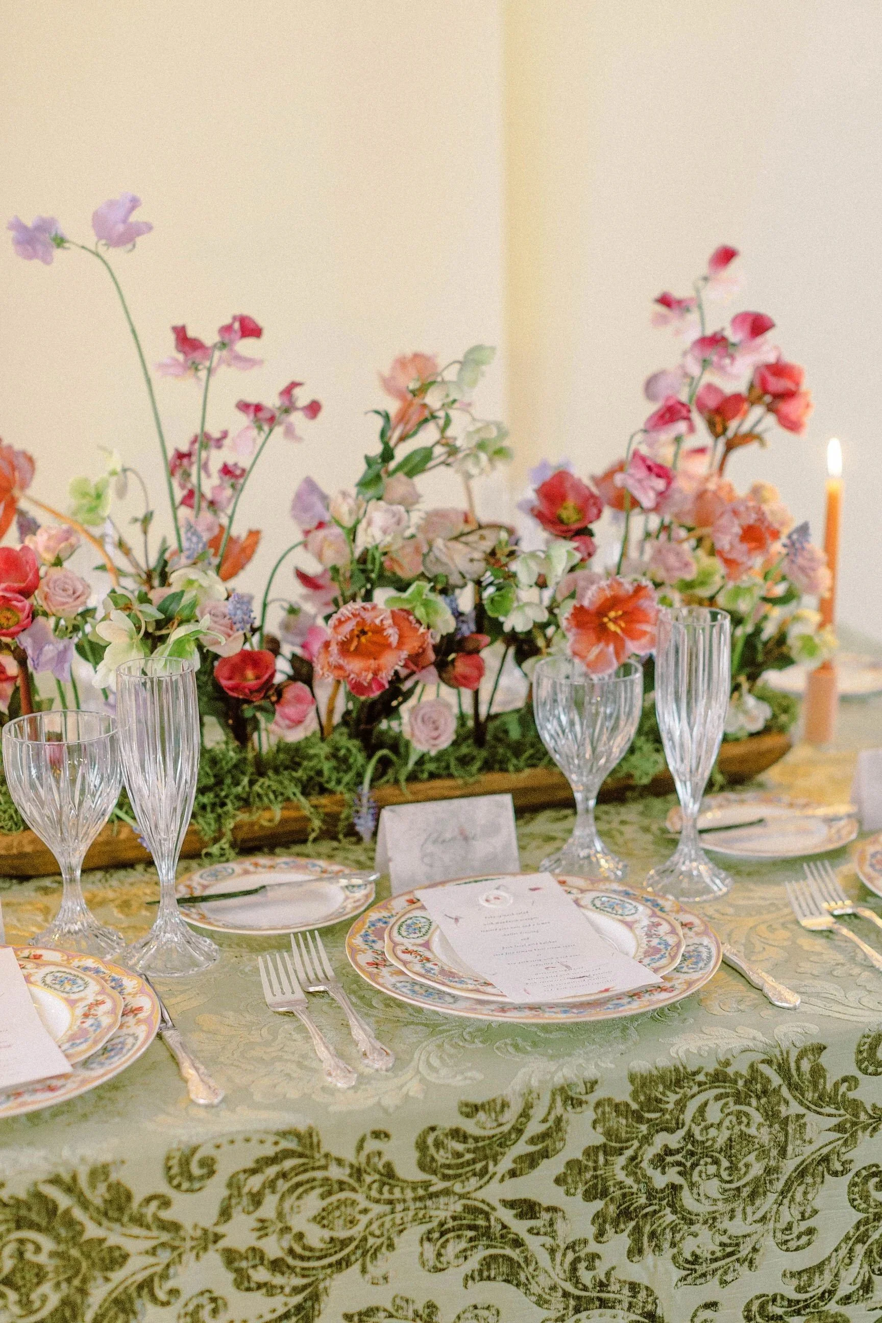 A table set for a formal event with a floral centerpiece, glassware, plates, silverware, and a menu card, all arranged on a green and gold patterned tablecloth, with candles in the background.