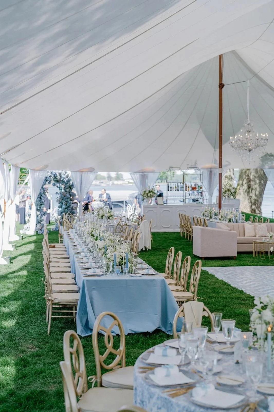 Elegant outdoor wedding reception setup under a large white tent with long dining tables and floral arrangements, overlooking a body of water.