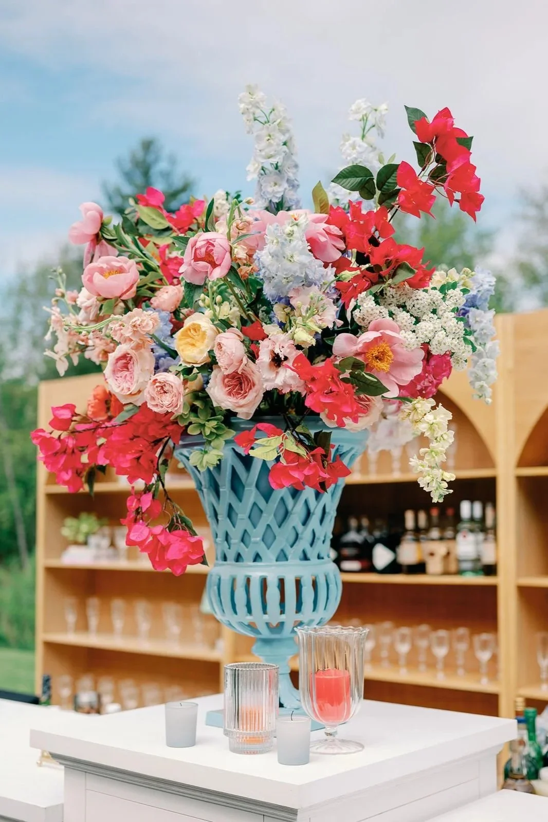 Large colorful wedding floral arrangement in a blue decorative vase on a white table, with candles and glasses around it, set outdoors with a wooden backdrop and a partly cloudy sky.