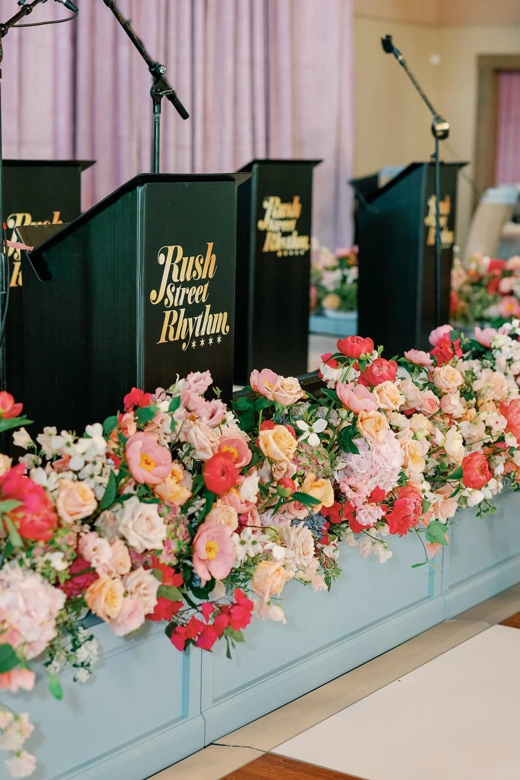 Mic stands behind black podiums labeled "Rush Street Rhythm" decorated with pink and white flowers at a stage
