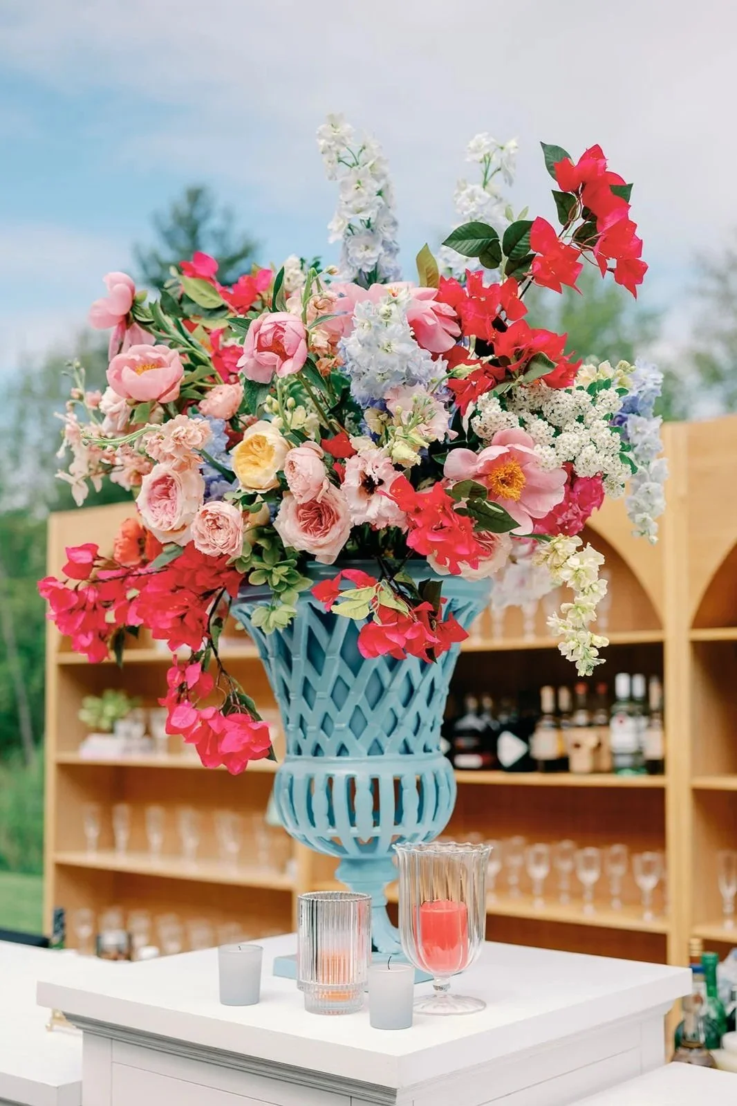 Large bouquet of pink, red, and white flowers in a blue decorative vase on a white table with candles, with a wooden shelving unit in the background outdoors.