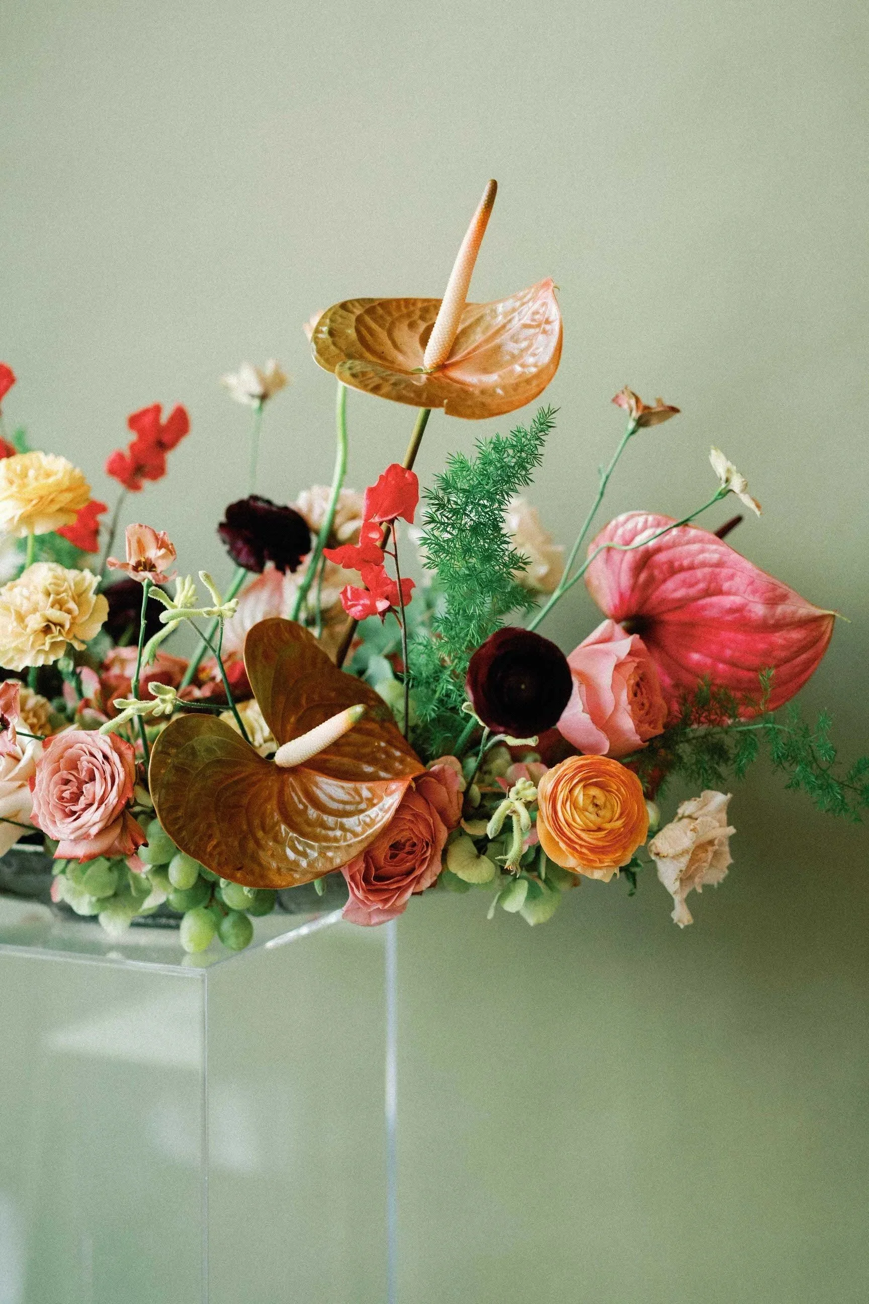 A floral arrangement with red, pink, yellow, and white flowers, large green leaves, and a prominent brown leaf with a cream-colored stem, set against a muted green background.