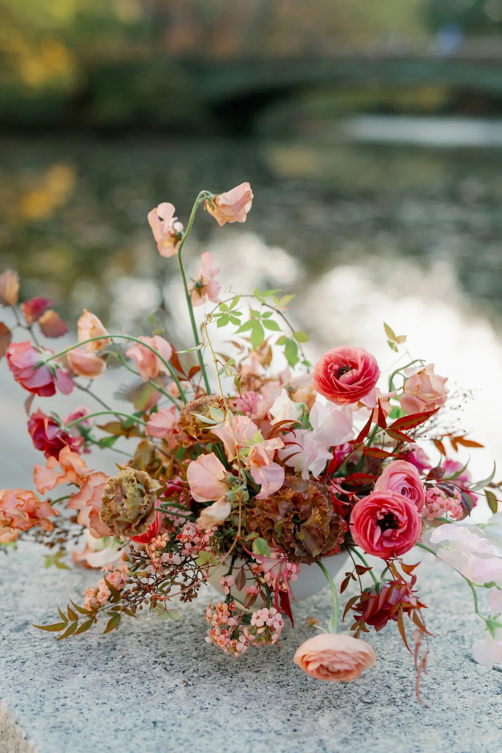 A bouquet of pink, red, and peach flowers, including roses and ranunculus, resting on a cement surface with a blurred outdoor background.