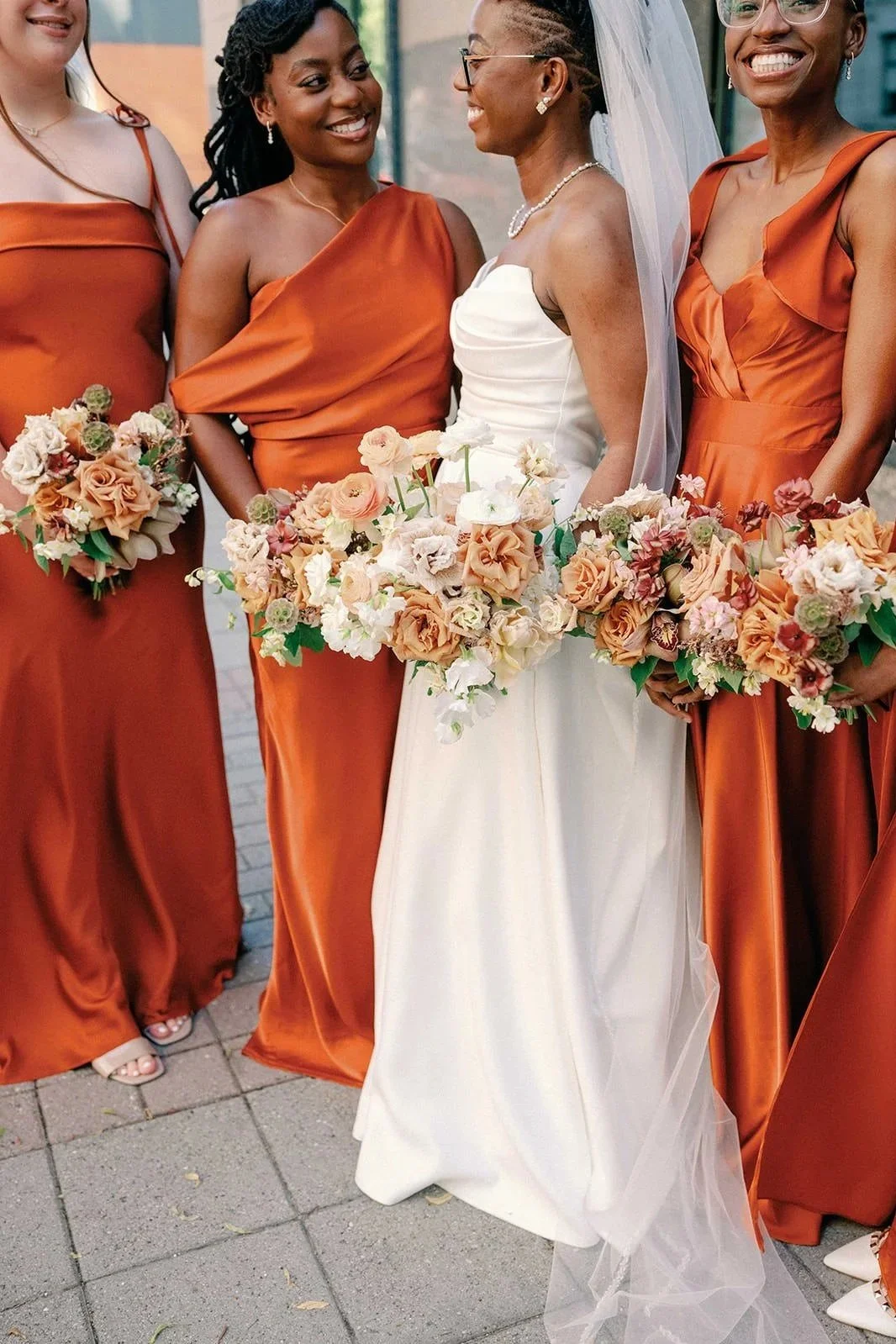 Wedding bride in a wedding dress, holding a bouquet of peach, cream, and white flowers, surrounded by bridesmaids in rust-colored dresses, each holding similar bouquets, on a paved outdoor setting.