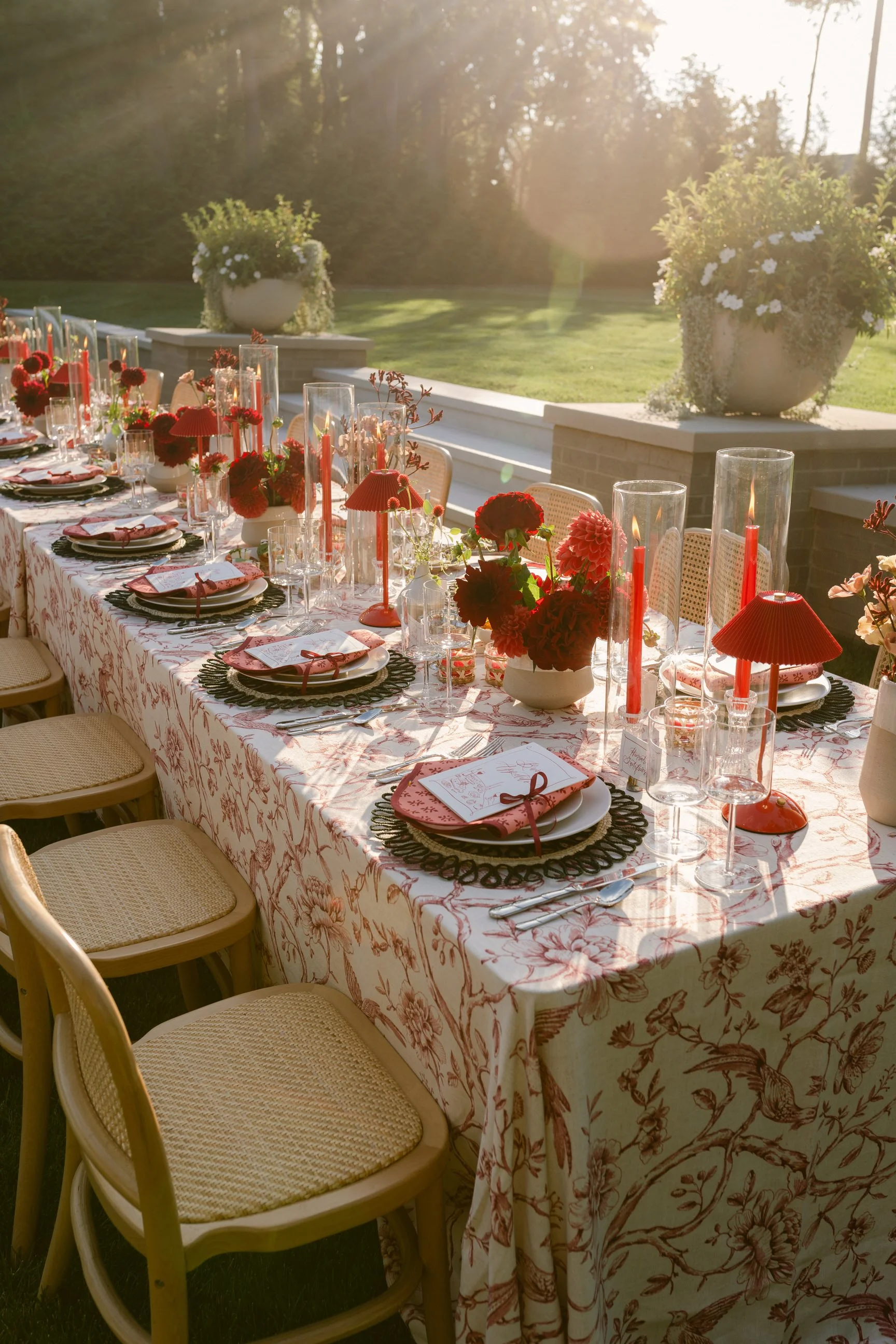 A long dining table outdoors set for a celebration with red and pink flowers, candles, and place settings, on a patio with large planters of white flowers and a grassy yard in the background, sun shining in Michigan.