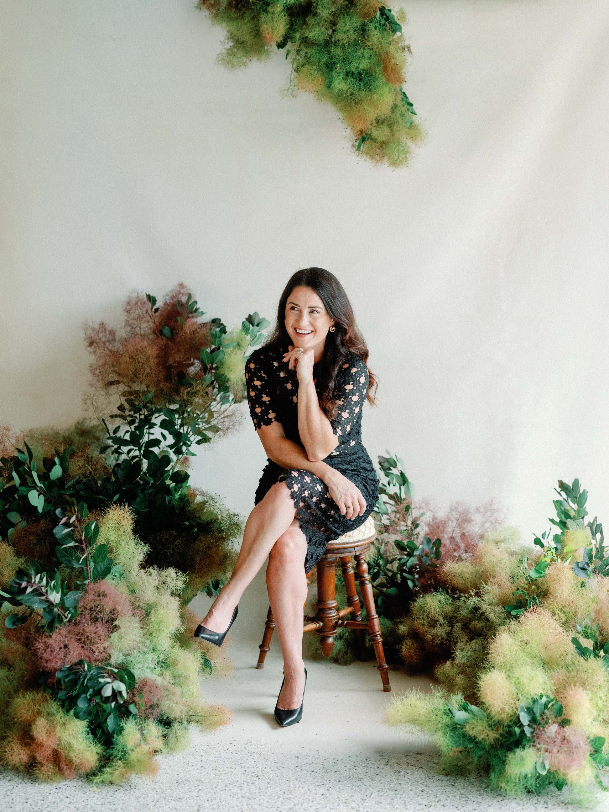 Rhiannon Bosse, Creative director of Rhiannon Bosse Celebrations sitting on wooden stool surrounded by smoke bush installation