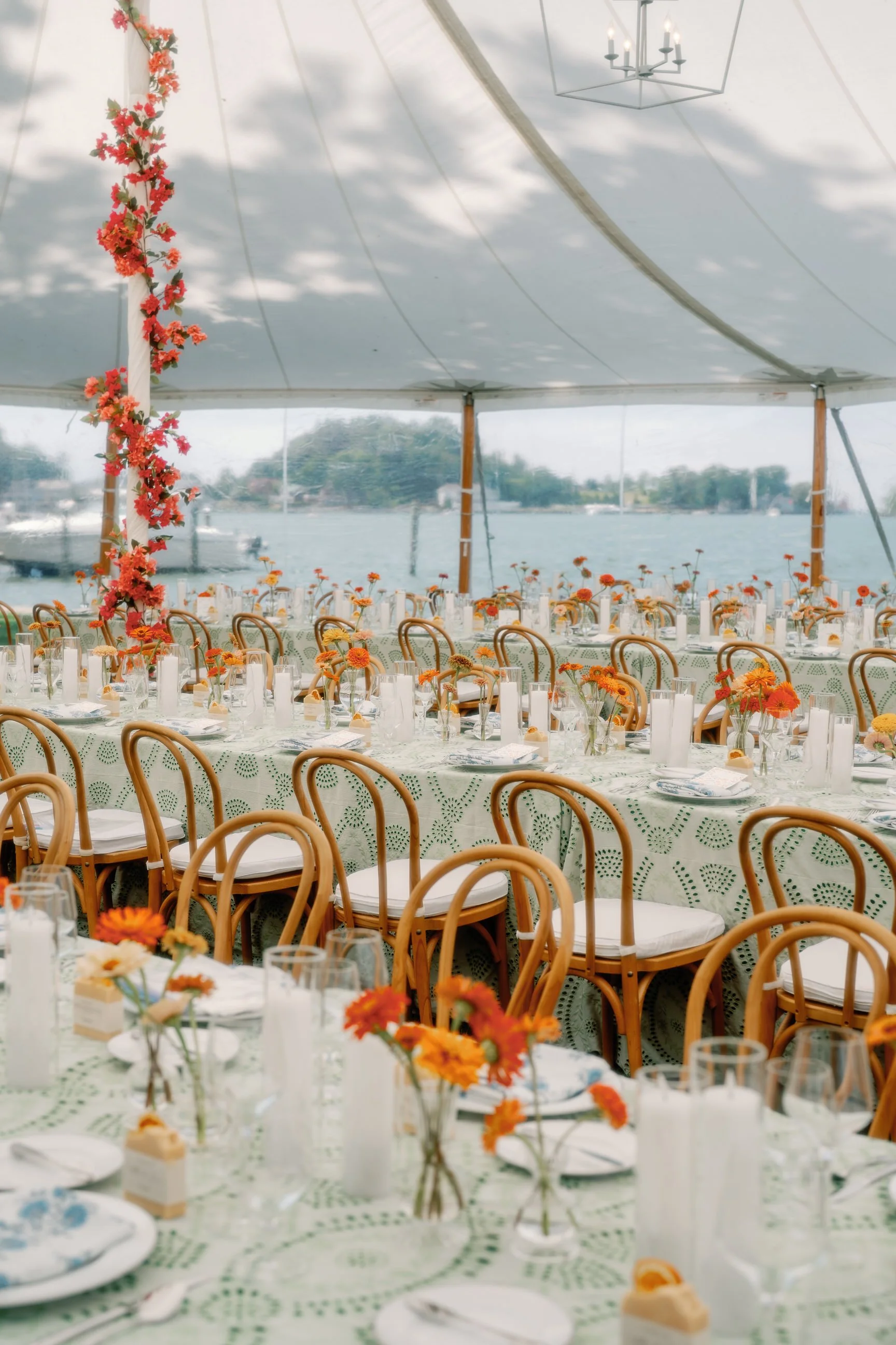 Long banquet tables with floral centerpieces, set for a formal wedding reception event under a large tent, overlooking a body of water.