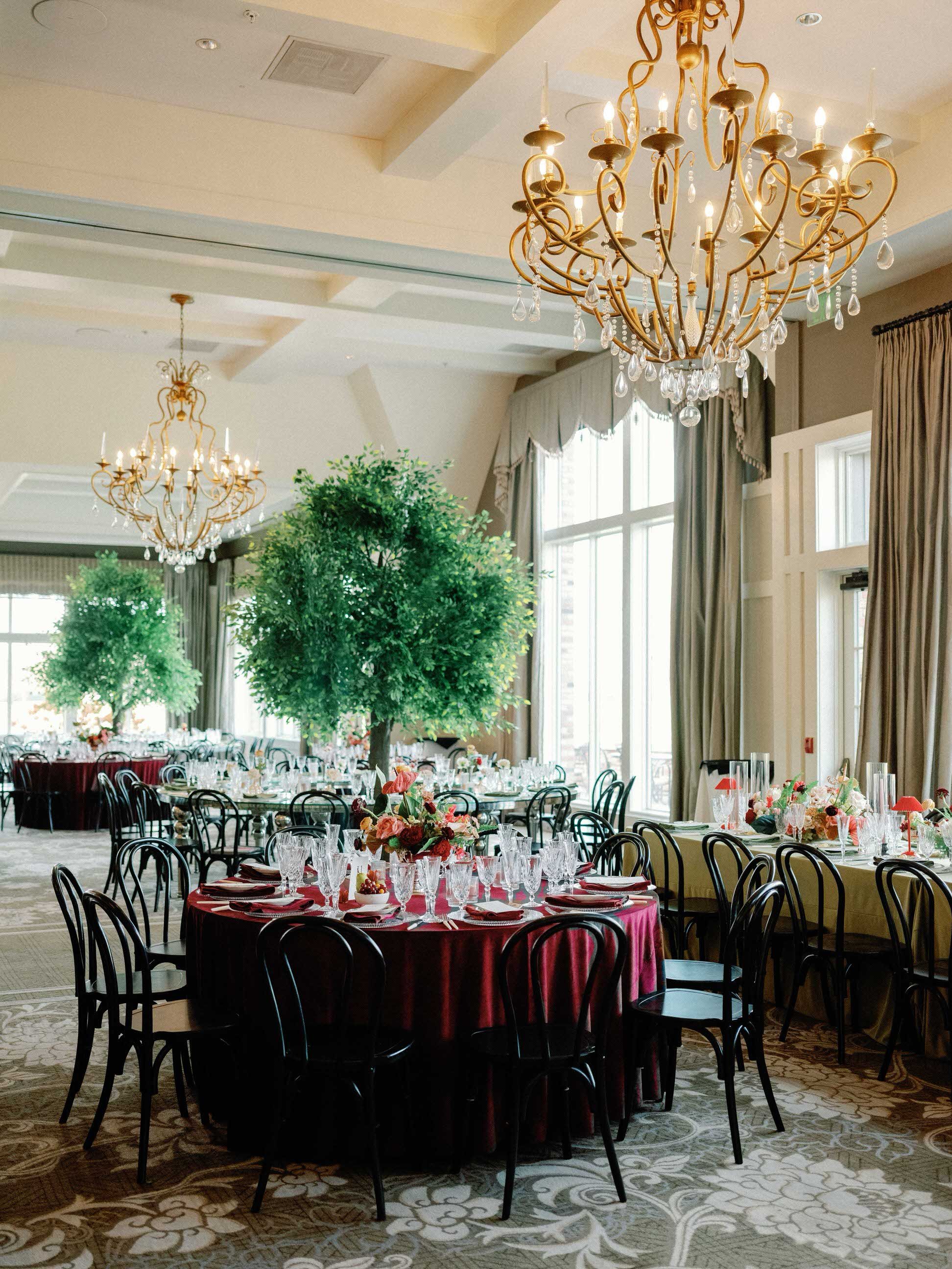 Elegant banquet hall decorated for a formal event with round tables draped in burgundy tablecloths, set with glassware and floral centerpieces, surrounded by black chairs, with large windows, chandeliers, and tall potted plants in the background.