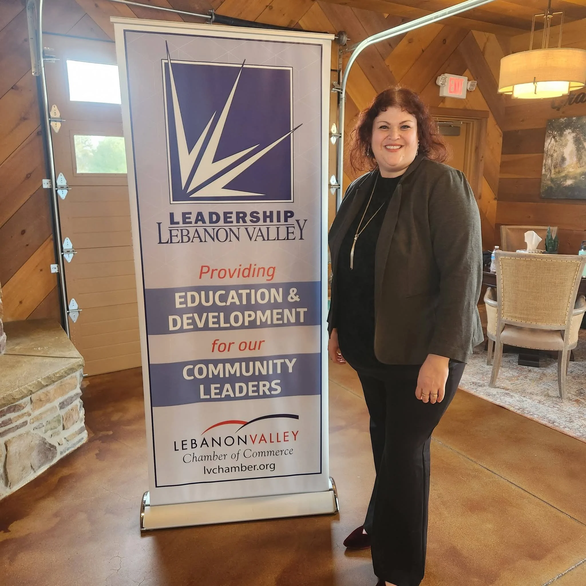 A professional woman standing next to a sign for Leadership Lebanon Valley, which includes the logo for the Lebanon Valley Chamber of Commerce. Hiring help and recruiting services by a female-owned, small business in Lebanon.