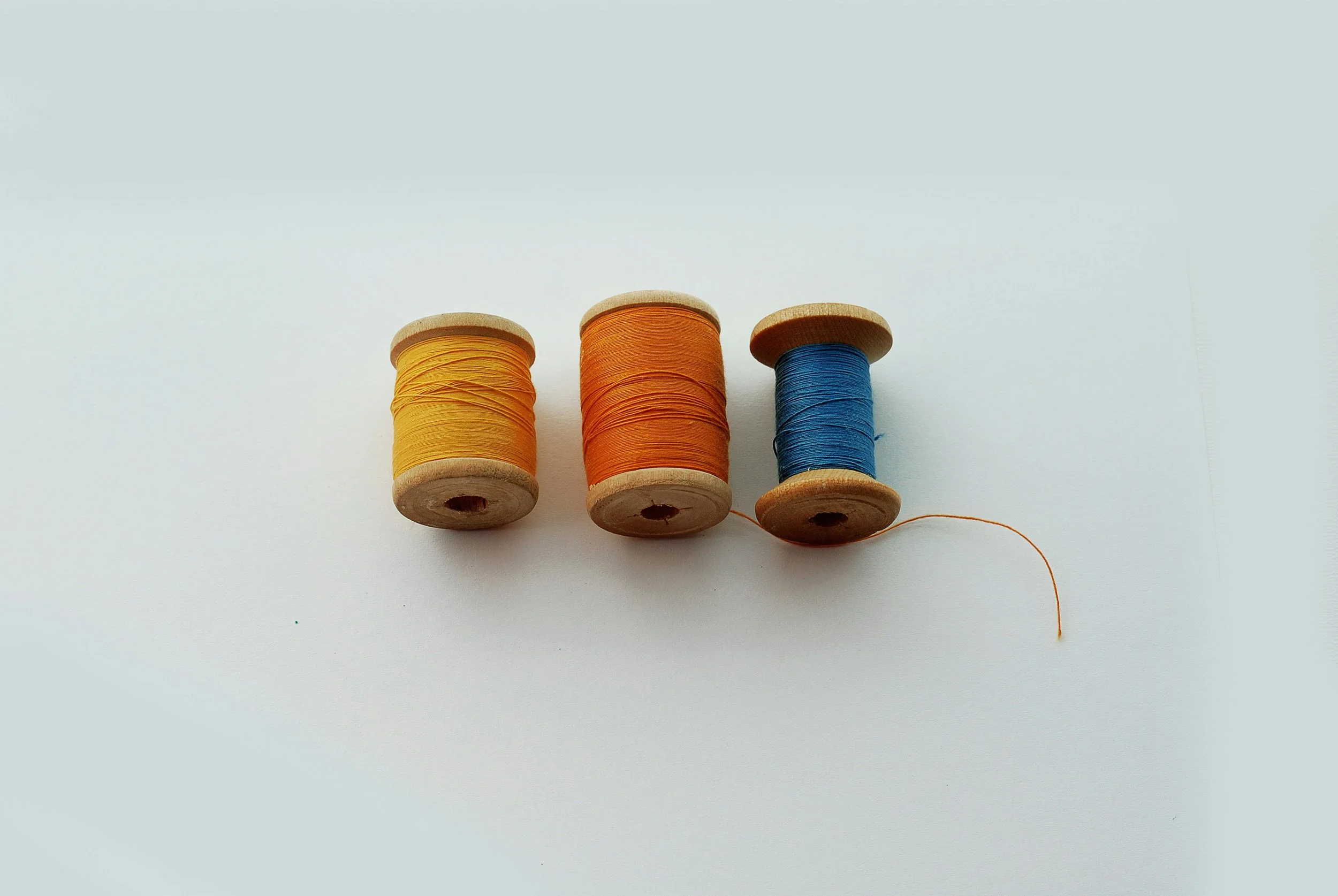 Three wooden spools of thread, from left to right: yellow, orange, and blue, placed on a white background.