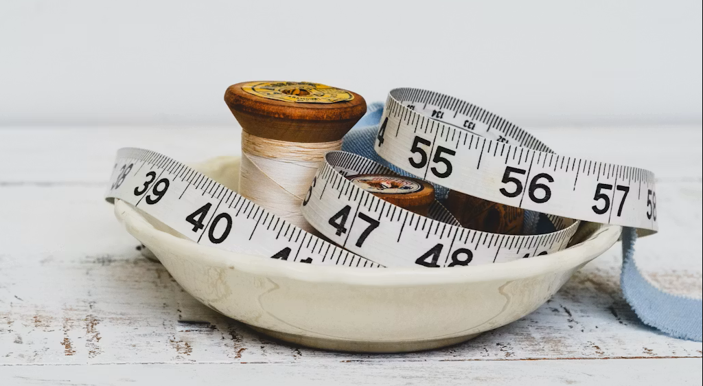 Sewing pins, a measuring tape, and a cloth in a ceramic dish on a wooden surface.