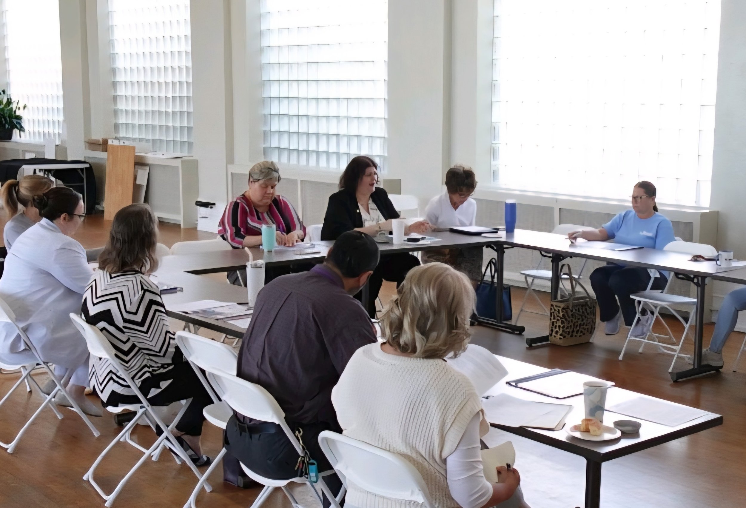 A diverse group of professional women and leaders seated around a table in a well-lit room with large windows, engaged in a meeting or discussion about hiring needs, skilled workers, and employment.