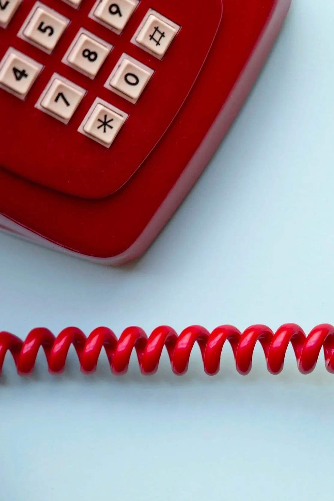 Close-up of a red corded telephone keypad and coiled handset cord on a light blue background.