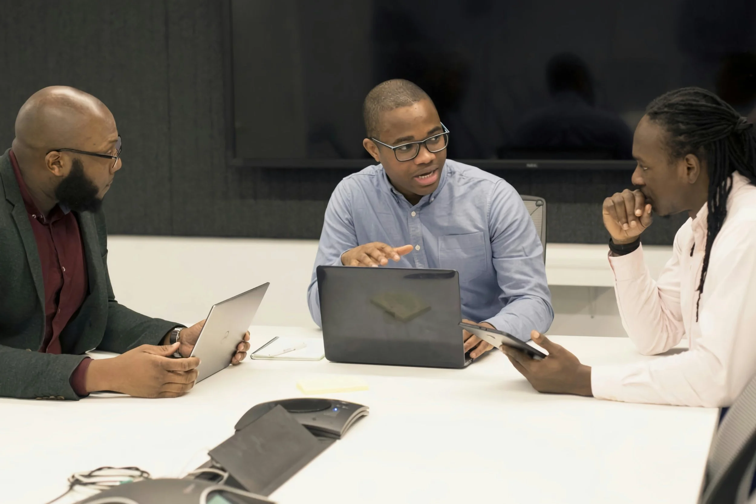 Three men engaged in a discussion at a conference table in a modern office environment, using laptops and tablets.