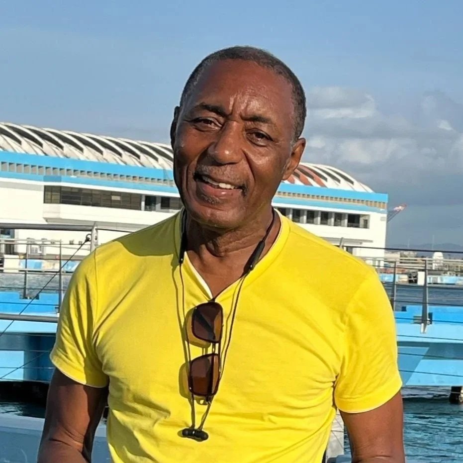 A smiling middle-aged man with dark skin, wearing a yellow t-shirt, sunglasses hanging from his neck, and earphones, standing outdoors near a body of water with a modern building in the background. HydroFLOW Jamaica Owner, Winston Hawthorne.
