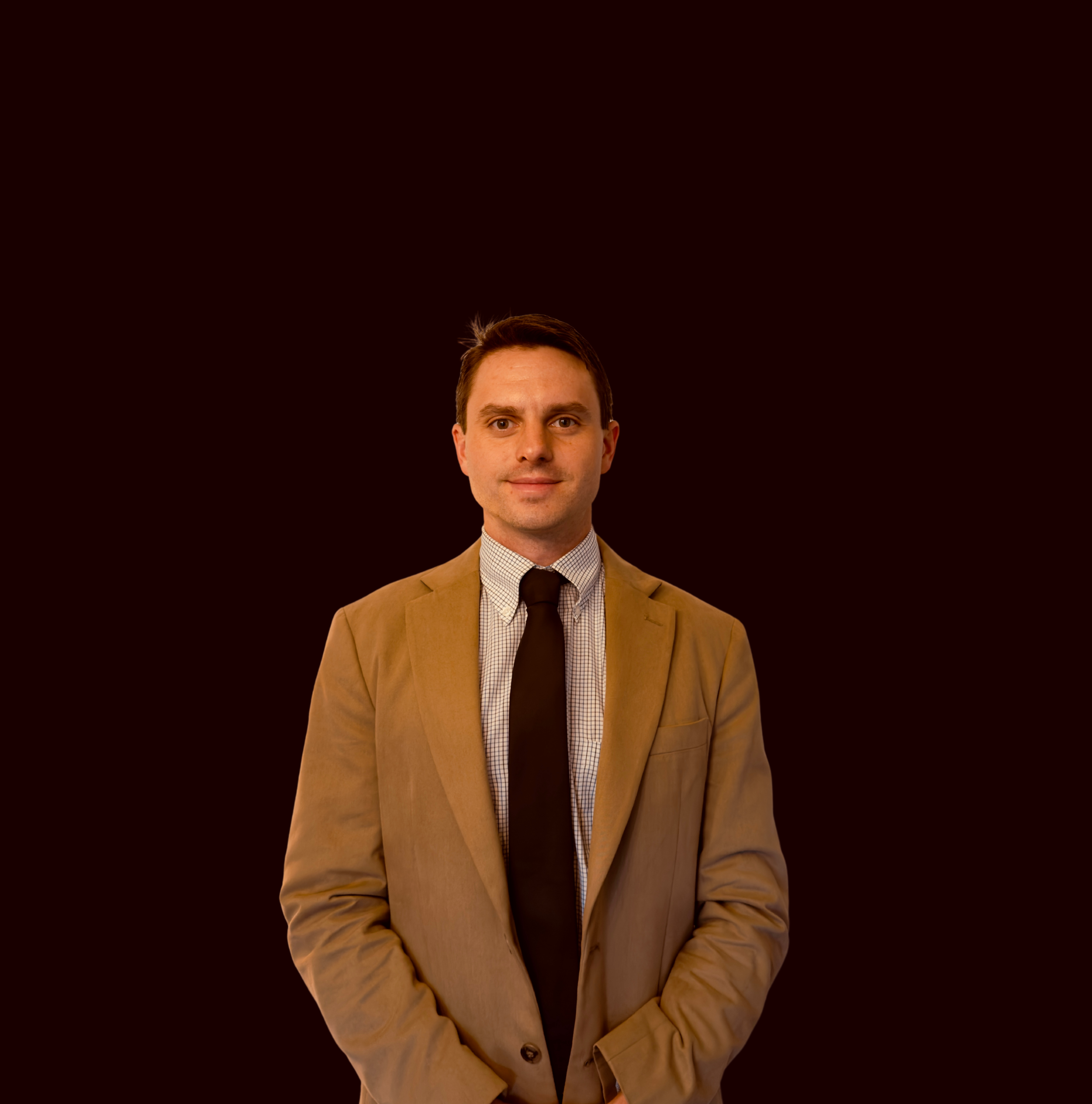 A man in a tan blazer, white checkered shirt, and black tie standing against a dark background.